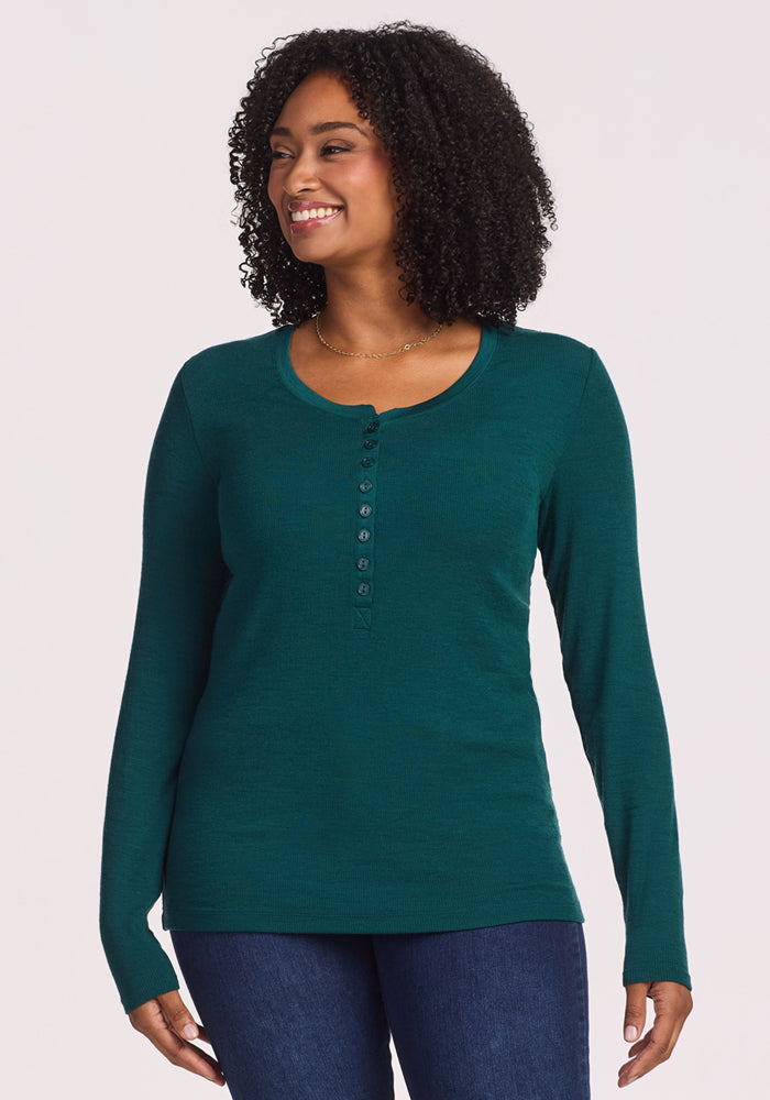 A woman with curly hair smiles while wearing the Woolx Reese Ribbed Henley in Riverbank Green and blue jeans, standing against a plain, light background. | Maysa is 5’3”, wearing a size M