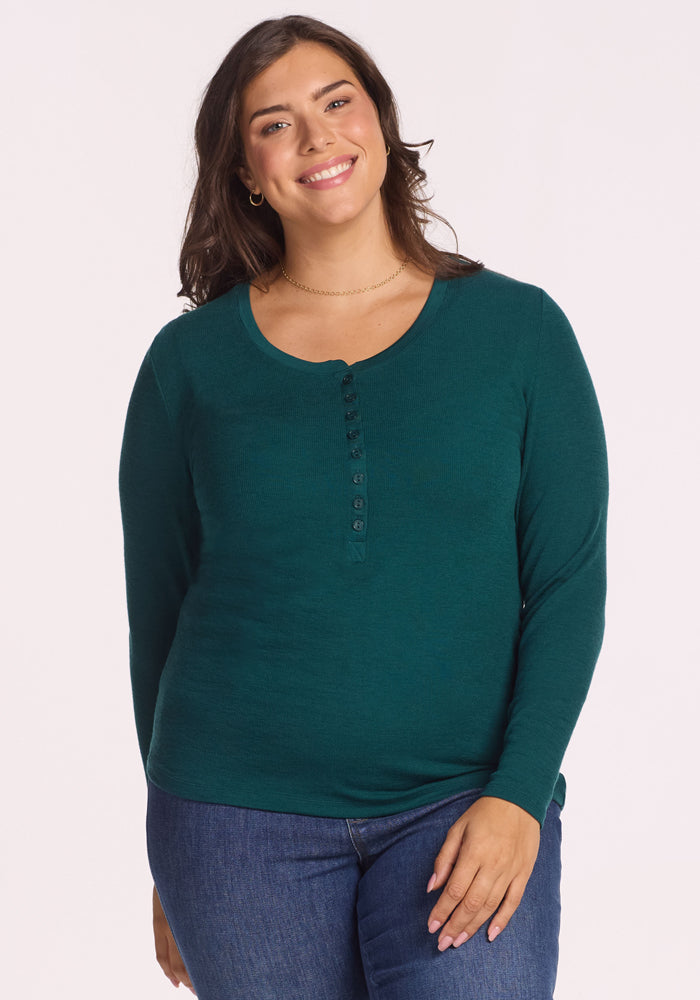 A woman with long brown hair wears the Woolx Reese Ribbed Henley in Riverbank Green and blue jeans, smiling as she stands against a plain, light-colored background. | Juliana is 5'10", wearing a size XL
