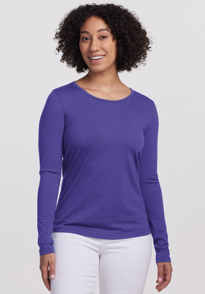 Smiling woman with curly hair wears Woolx Remi Long Sleeve T-Shirt in Berry Indigo and white pants against a plain background. | Tori is 5'7", wearing a size S