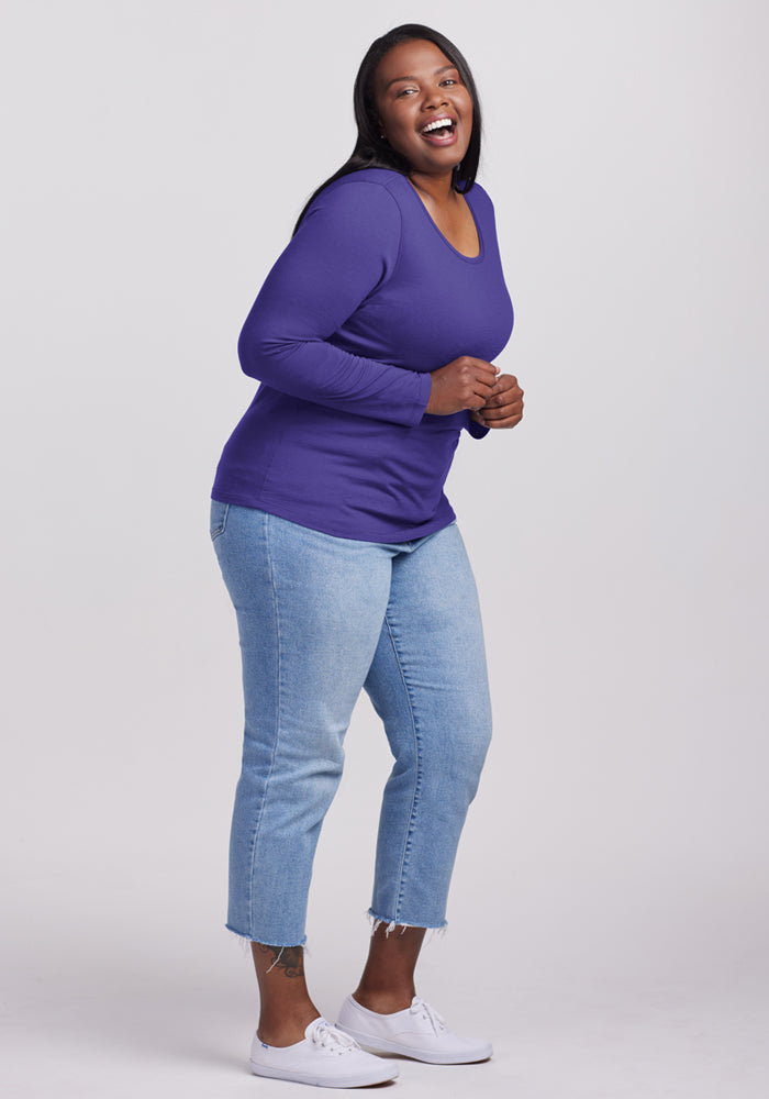 Woman in Woolx Remi Long Sleeve T-Shirt - Berry Indigo, light blue jeans, and white sneakers stands against a light background.