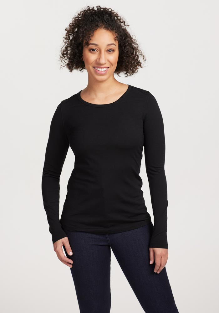 A person with curly hair is smiling and posing in a Woolx Remi Long Sleeve T-Shirt OLD in black, paired with dark pants against a plain backdrop.