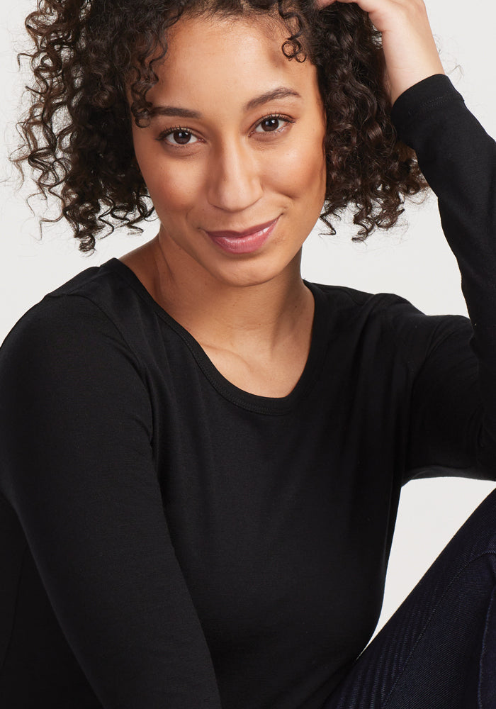 A person with curly hair smiles softly while resting their hand on their head, wearing a Remi Long Sleeve T-Shirt in black from Woolx. They sit against a light background, enjoying the comfort and odor-resistant properties of the Merino wool fabric.