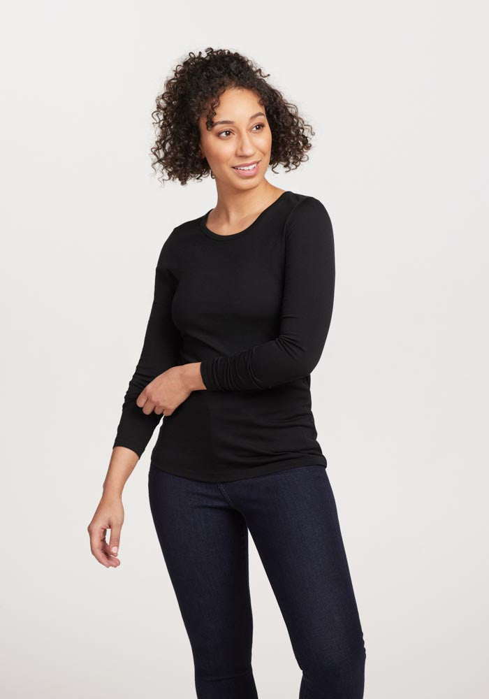 A person with curly hair is wearing a fitted black Remi Long Sleeve T-Shirt by Woolx, made of lightweight fabric, paired with dark jeans. They are standing against a plain light background, smiling and looking slightly to the side.