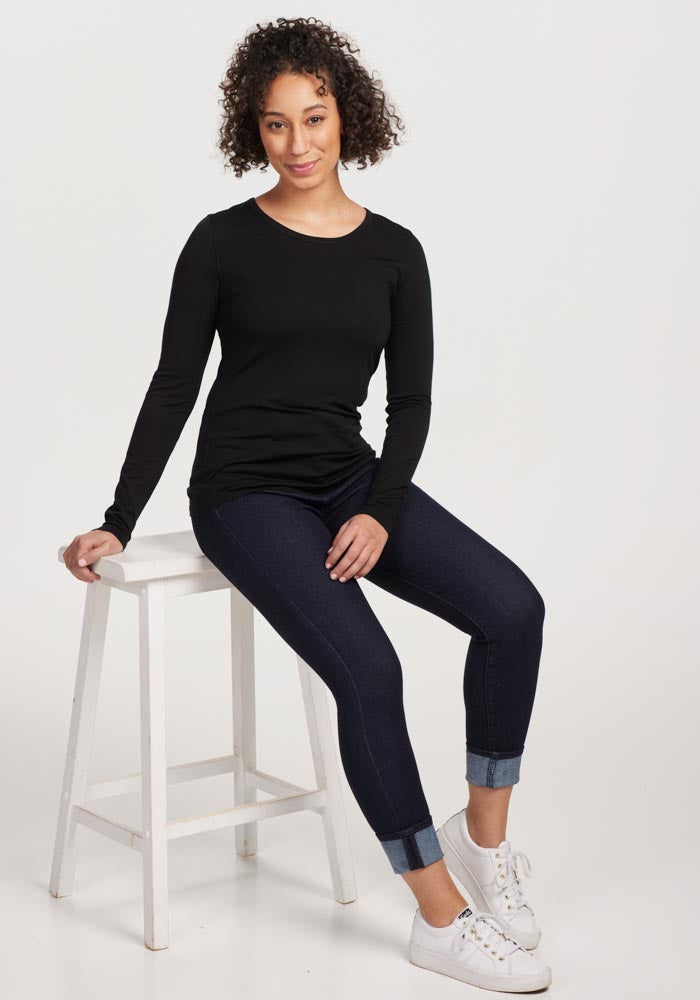 A person with curly hair sits on a white stool, dressed in a black Woolx Remi Long Sleeve T-Shirt made of lightweight fabric, dark rolled-up jeans, and white sneakers. The background is plain white.
