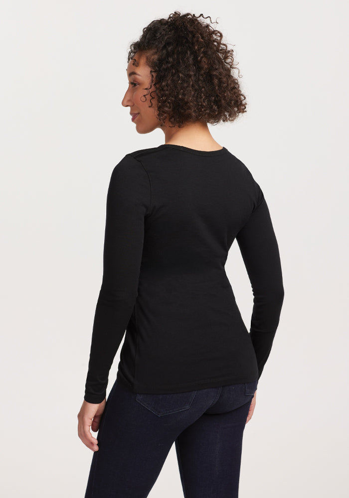 A woman with curly hair stands sideways, facing right, wearing the Remi Long Sleeve T-Shirt - Black by Woolx and dark jeans. The background is plain white.