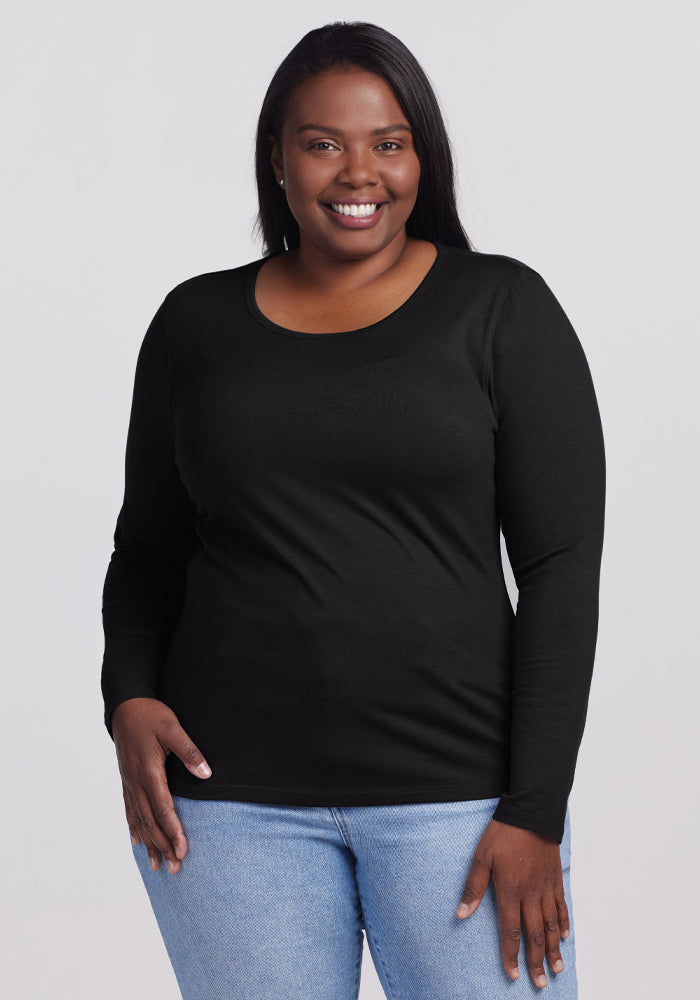 A person with long dark hair smiles warmly at the camera, wearing a Woolx Remi Long Sleeve T-Shirt in black and light blue jeans. The cozy Merino wool top complements their look as they stand against a plain light gray background. | Le'Quita is 5'11", wearing a size XL