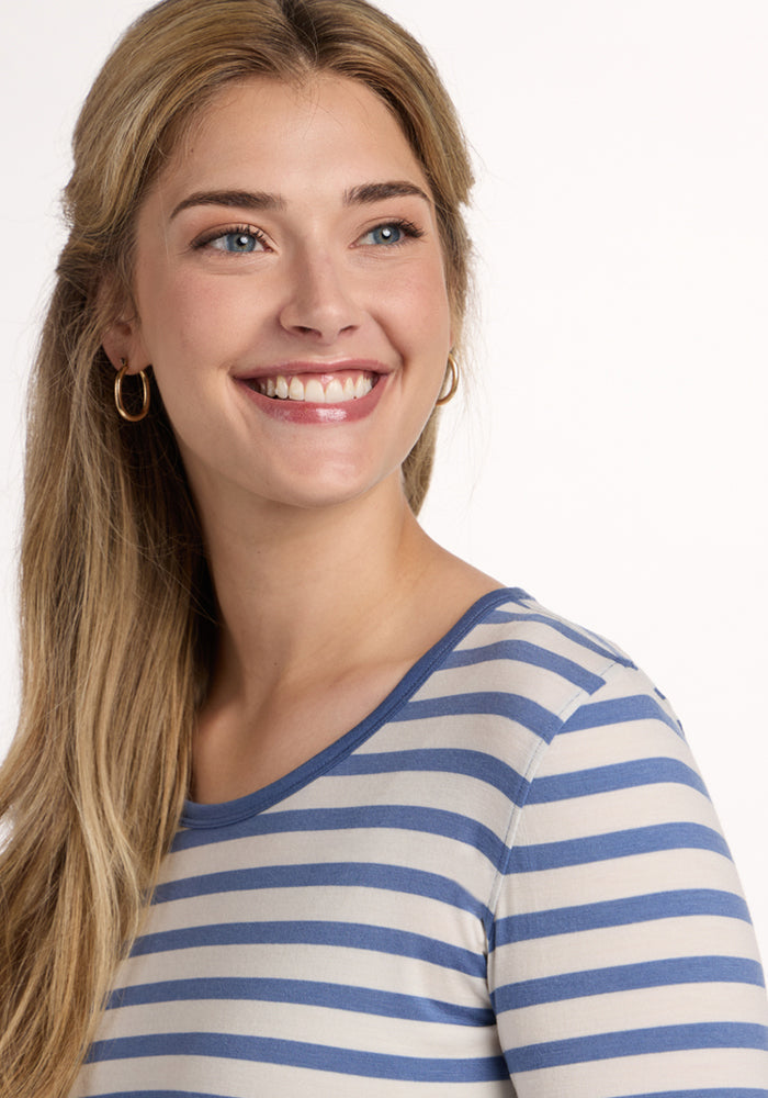A woman with long blonde hair and gold hoop earrings smiles while wearing the Woolx Remi Long Sleeve T-Shirt in Blue Sand Stripe, looking slightly to the side against a plain background.