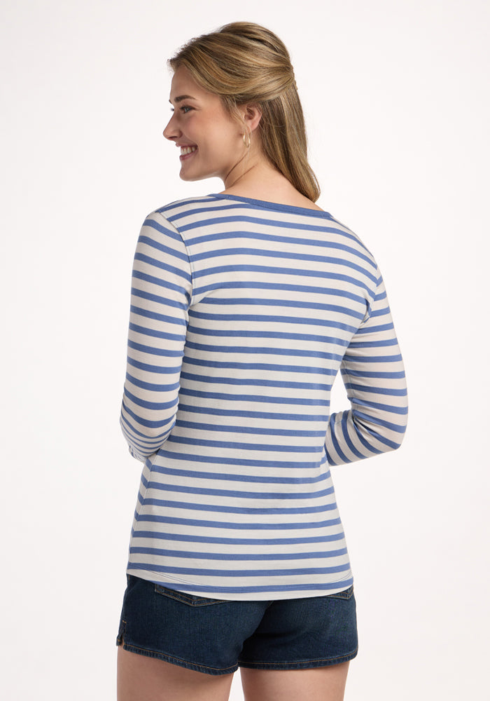 A blonde woman smiles while facing away from the camera, wearing the Woolx Remi Long Sleeve T-Shirt in Blue Sand Stripe with dark denim shorts against a plain light background.