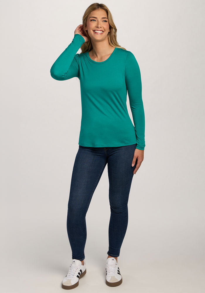 Woman smiles in a Woolx Remi Long Sleeve T-Shirt - Cabana Crush, blue jeans, and white sneakers against a plain background.