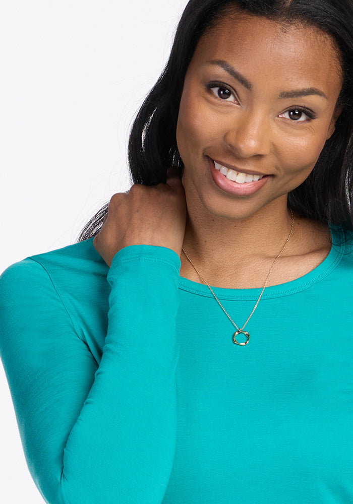 Smiling woman in a wrinkle-resistant Woolx Remi Long Sleeve T-Shirt in Cool Jade, gold necklace, touches her shoulder.