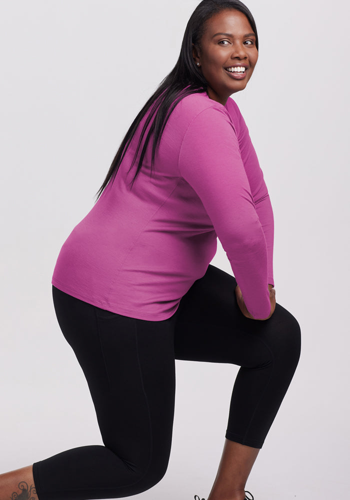 Smiling woman kneels in Woolx Remi Long Sleeve T-Shirt in Purple Wine and black leggings against a light background.