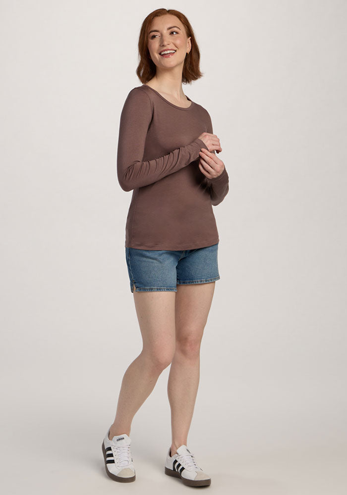 Woman smiling in Woolx Remi Long Sleeve T-Shirt - Peppercorn, denim shorts, and white sneakers against a plain background.