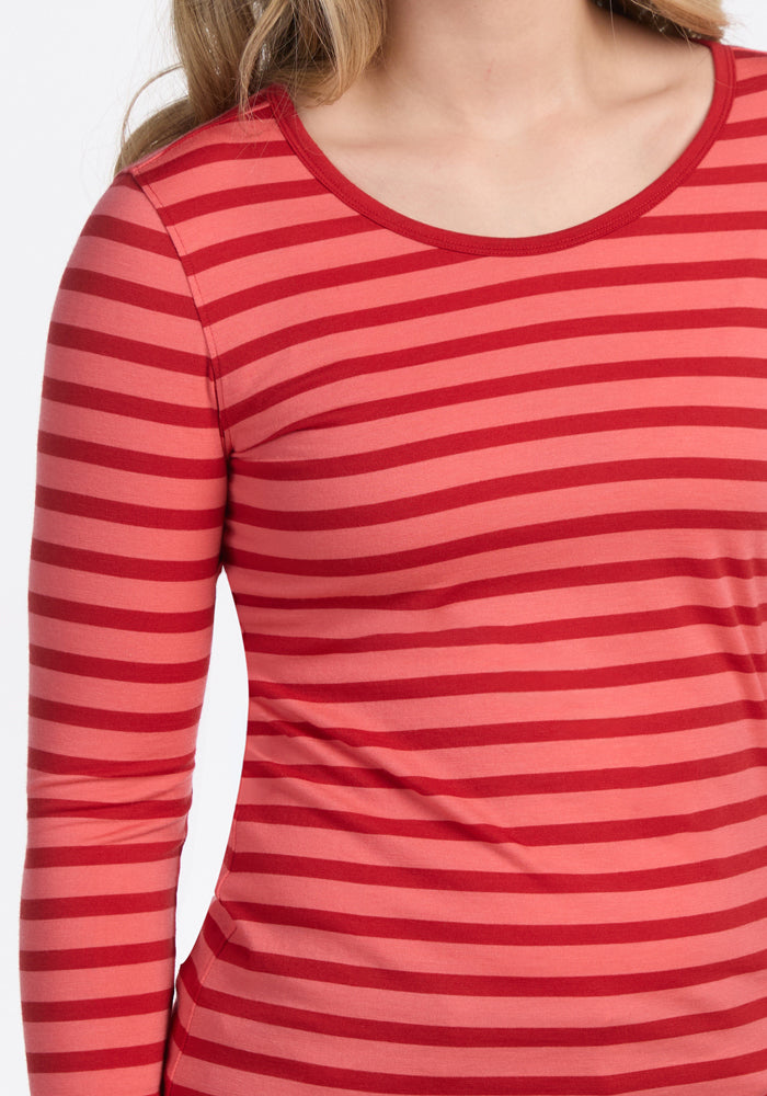 A person with light brown hair wears the Woolx Remi Long Sleeve T-Shirt in Wildberry Stripe, featuring horizontal red and pink stripes. The image shows their upper body from shoulders to waist.