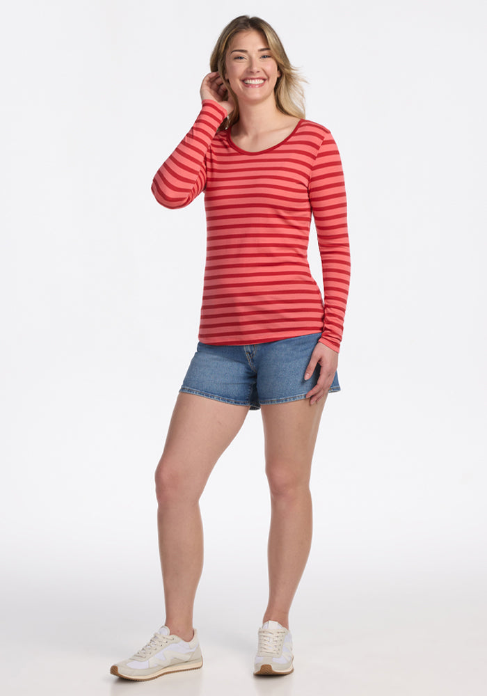 A person with light brown hair smiles, touching their head, wearing the Woolx Remi Long Sleeve T-Shirt in Wildberry Stripe, denim shorts, and white sneakers against a plain white background.