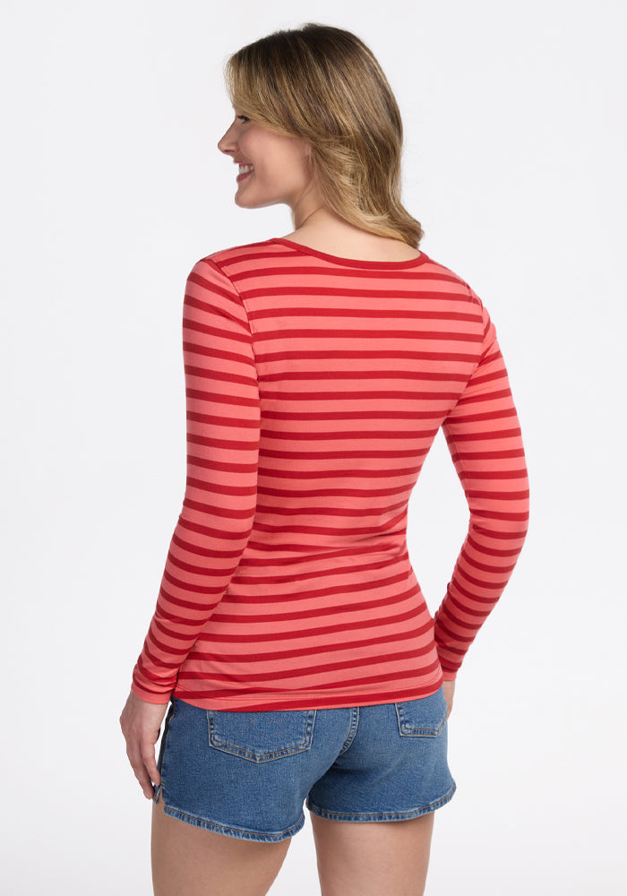 A woman smiles with her body turned slightly to the side, wearing the Woolx Remi Long Sleeve T-Shirt in Wildberry Stripe and denim shorts, set against a plain white background.