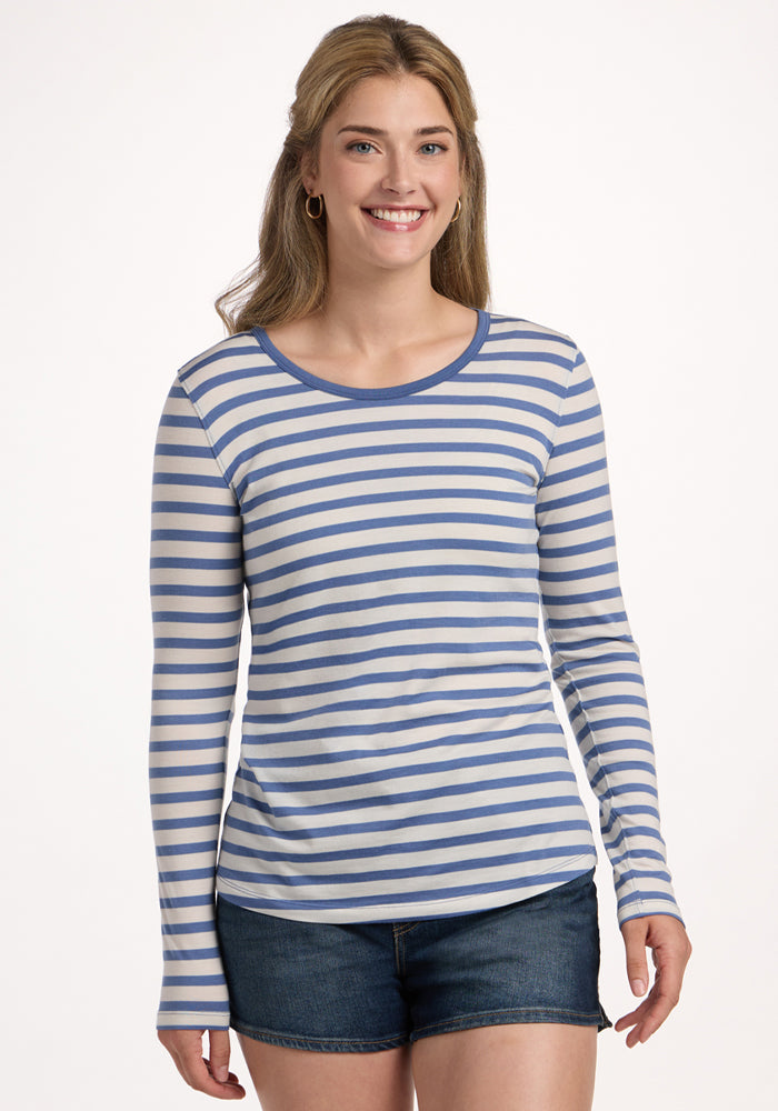 A smiling woman with long, light brown hair wears the Woolx Remi Long Sleeve T-Shirt in Blue Sand Stripe and denim shorts, standing against a plain white background. | Ariana is 5'11", wearing a size S