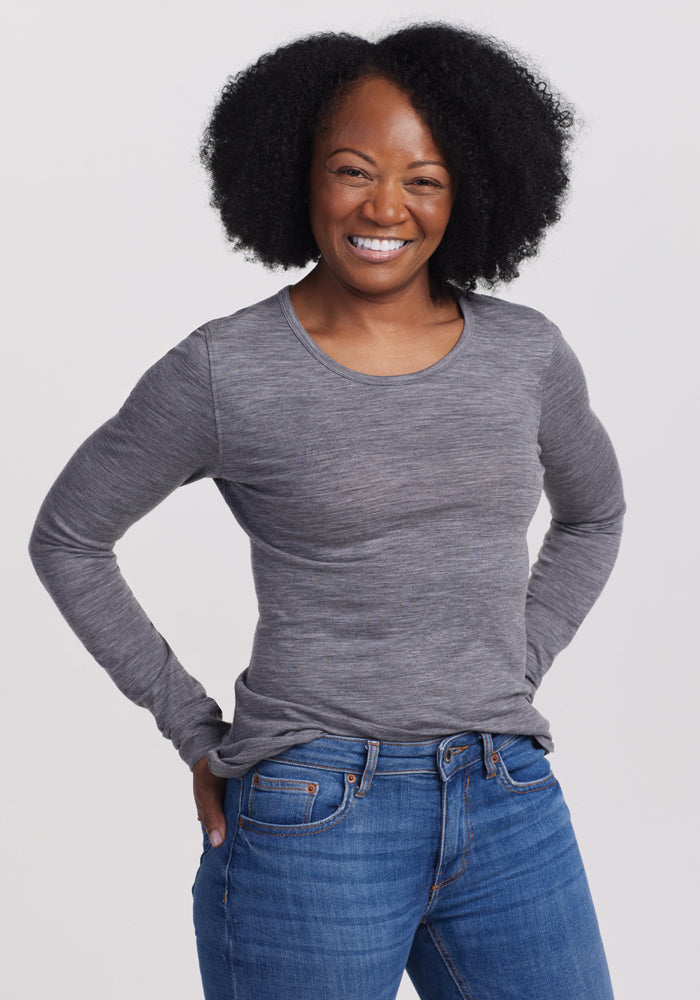 A curly-haired person in a Woolx Remi Long Sleeve T-Shirt - Graphite Heather and blue jeans smiles with hands on hips against a plain white background. | Pechaz is 5’4”, wearing a size S