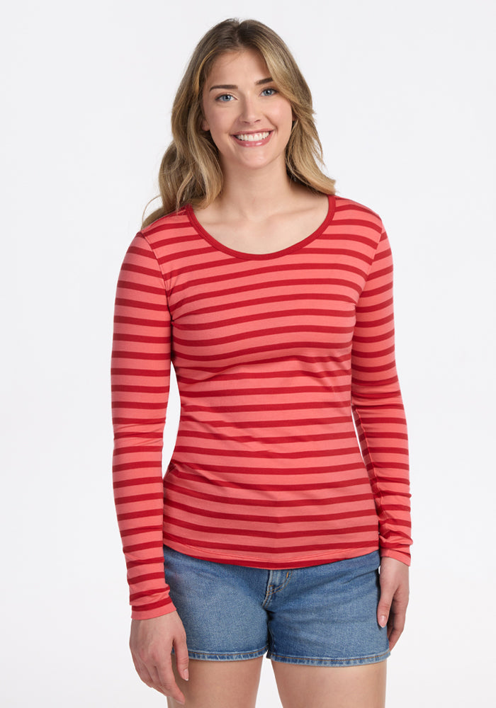 A woman with long light brown hair smiles against a white background, wearing the Woolx Remi Long Sleeve T-Shirt in Wildberry Stripe paired with denim shorts. | Ariana is 5'11", wearing a size S