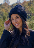 A woman with long dark hair smiles outdoors among green trees, wearing the Woolx Ria Chunky Beanie in black and a black top, with a blurred background of foliage and blue sky.