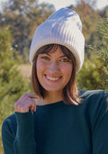 A young woman smiles outdoors in soft sunlight, surrounded by green trees, wearing the Woolx Ria Chunky Beanie in Buttercream and a green sweater.
