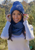 A woman smiles outdoors in a wooded area on a sunny day, wearing the Woolx Ria Chunky Beanie in Starry Night and a matching blue scarf with a white long-sleeve shirt.