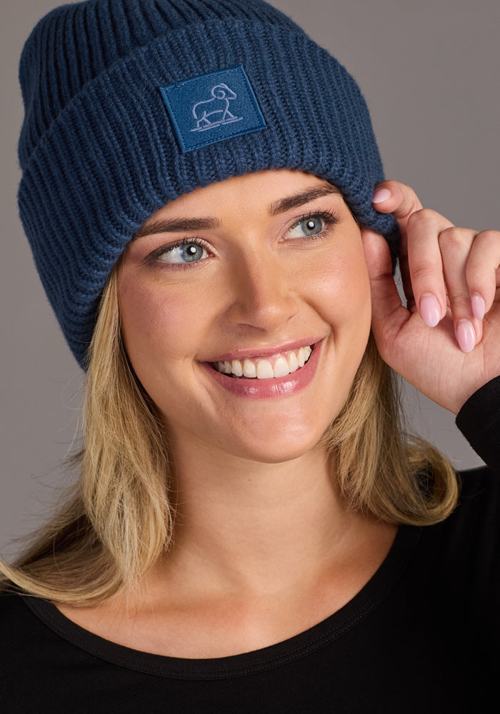 A woman smiles as she wears the Woolx Ria Chunky Beanie - Starry Night, featuring a white ram patch. Paired with a black top, she poses against a plain gray background, gently touching her cozy blue winter hat.