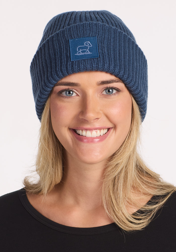 A smiling blonde woman wears the Woolx Ria Chunky Beanie in Starry Night, featuring a simple dog patch. She pairs it with a black top against a white background, highlighting this cozy winter accessory.