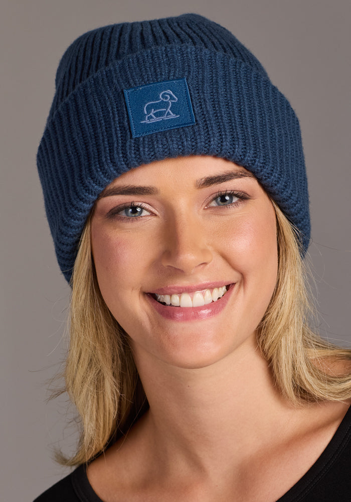 A smiling person with blonde hair wears the Woolx Ria Chunky Beanie - Starry Night, which features a patch with a simple animal outline, along with a black top, posing against a plain gray background.