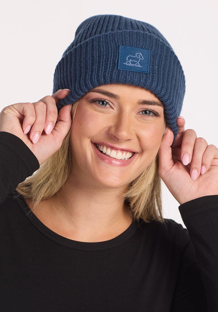 A smiling blonde person in a black top wears the Woolx Ria Chunky Beanie - Starry Night, a blue knit hat with a small patch of a simple white dog illustration, holding the cozy winter accessory by its edges.