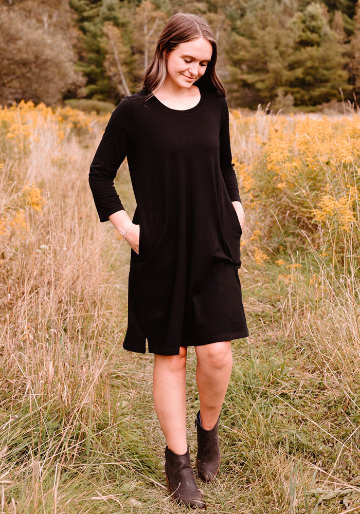 Woman in a Woolx Rissa A Line Dress - Black and boots stands in a grassy field with yellow wildflowers and trees.