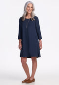 An older woman with long gray hair smiles against a white background, wearing the Woolx Rissa A Line Dress in Deep Navy with three-quarter sleeves, a long necklace, and brown loafers—perfect for fall-winter layering. | Tam is 5’8.5”, wearing a size S