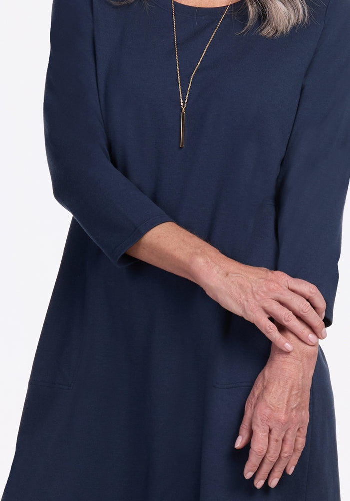 A woman wears the Woolx Rissa A Line Dress in Deep Navy with a gold pendant necklace, arms crossed and only her torso and arms visible—perfect for fall-winter layering.