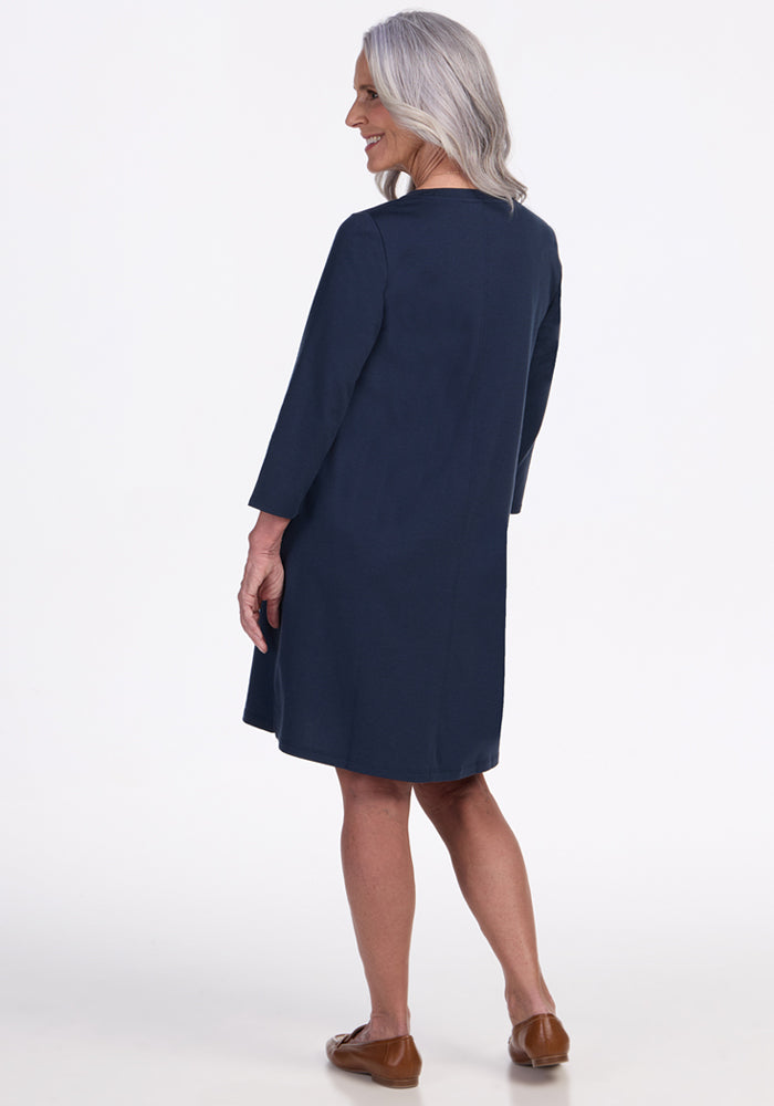 An older woman with long gray hair wears the Woolx Rissa A Line Dress in Deep Navy and brown shoes, standing facing away, looking over her shoulder and smiling—ideal for fall winter layering against a plain white background.