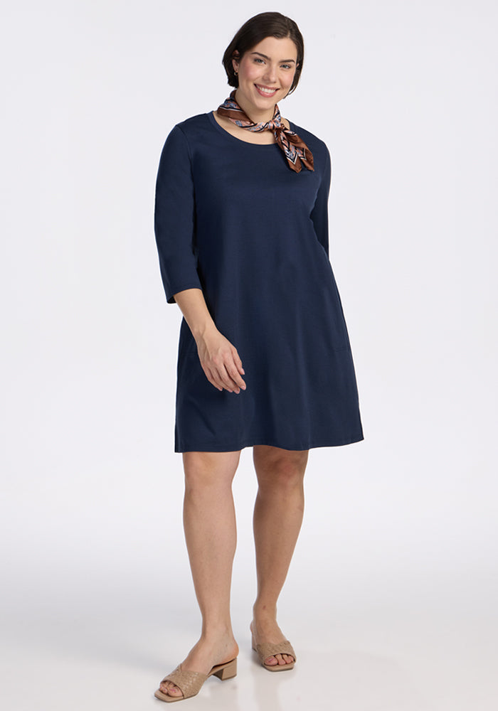 A woman models the Woolx Rissa A Line Dress in Deep Navy—knee-length Merino wool with three-quarter sleeves—styled with a patterned scarf and beige heeled sandals, ideal for fall/winter layering or as a chic travel dress. | Juliana is 5’10”, wearing a size XL