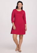 A woman with long dark hair smiles in the Woolx Rissa A Line Dress - Poppy Spark, a knee-length, bright red Merino wool dress with three-quarter sleeves and beige flats—ideal for fall/winter layering against a plain light background. | Sarah is 5'8", wearing a size S