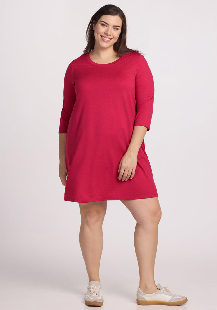 A woman with long brown hair smiles in the Woolx Rissa A Line Dress - Poppy Spark, a knee-length, bright pink Merino wool dress with three-quarter sleeves and white sneakers, set against a plain light background. | Juliana is 5'10", wearing a size XL
