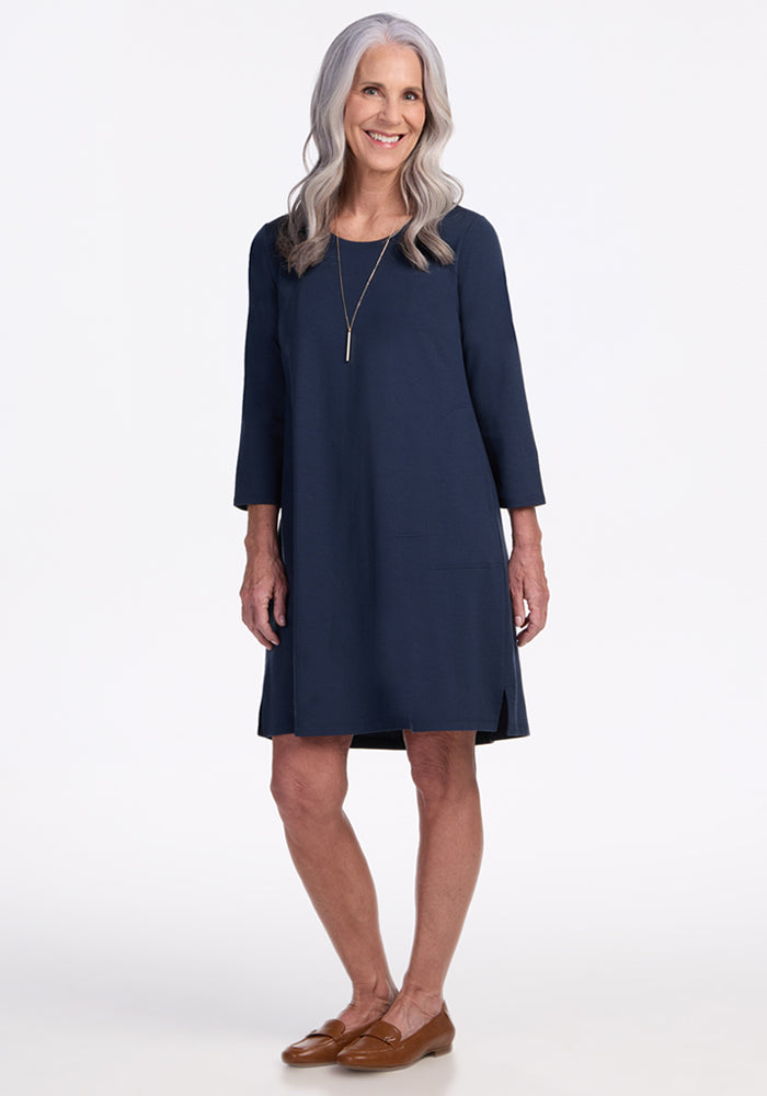 An older woman with long gray hair smiles against a white background, wearing the Woolx Rissa A Line Dress in Deep Navy with three-quarter sleeves, a long necklace, and brown loafers—perfect for fall-winter layering. | Tam is 5’8.5”, wearing a size S