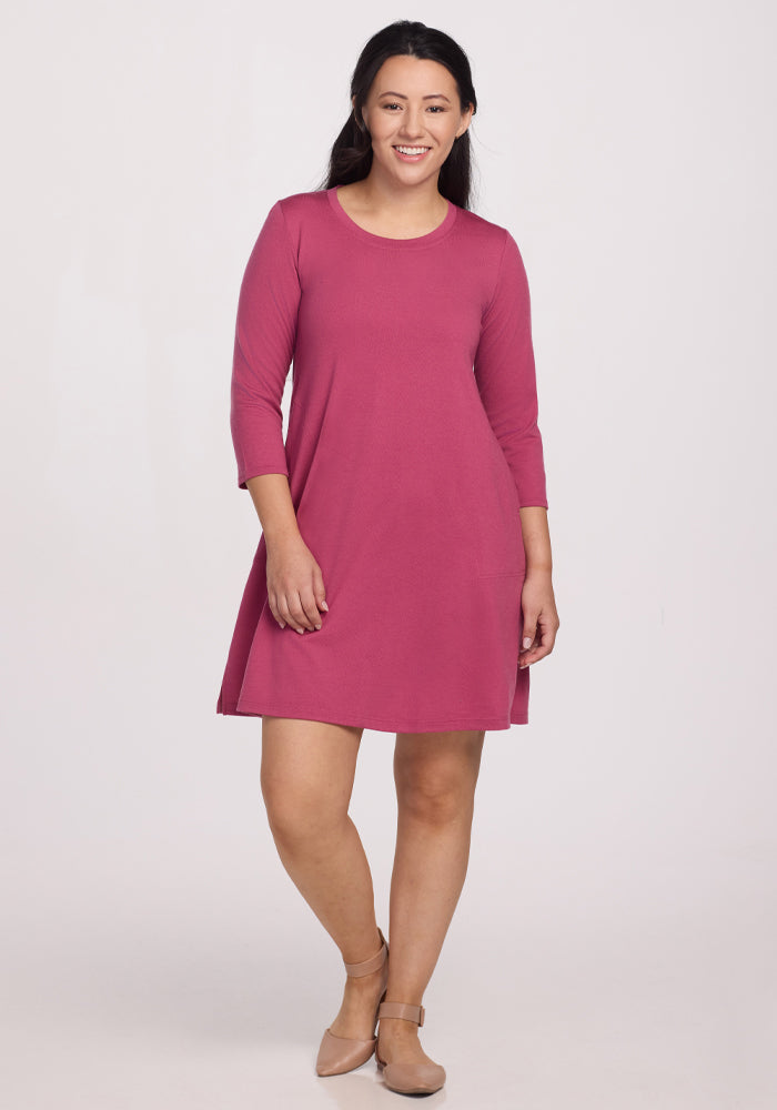 A woman with long dark hair wears the Woolx Rissa A Line Dress in Mulberry, smiling and standing against a plain white background.
