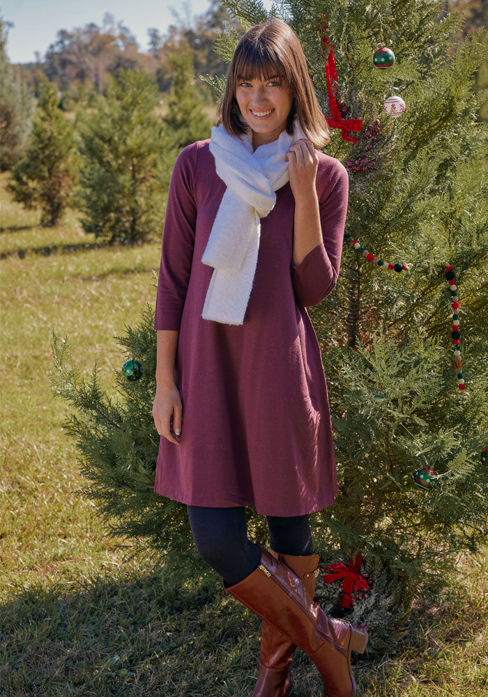 A woman wearing the Woolx Rissa A Line Dress in Toasted Garnet, paired with black leggings, brown boots, and a white scarf, smiles beside an outdoor Christmas tree—an ideal fall and winter layering look.