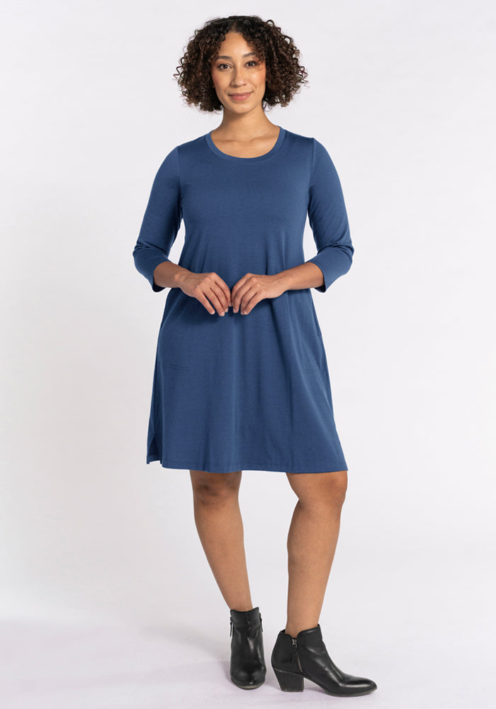 A woman with curly hair smiles gently, standing in a knee-length, long-sleeve Woolx Rissa A Line Dress in Starry Night and black ankle boots. She clasps her hands in front against a plain white background—ideal for fall and winter layering. Final sale.