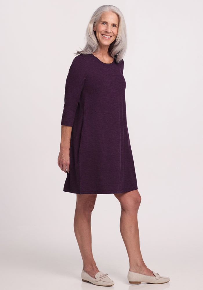 A smiling woman with long gray hair wears the Woolx Rissa A Line Dress in Deep Plum, a knee-length Merino wool piece perfect for fall and winter layering, paired with light slip-on shoes against a plain white background.