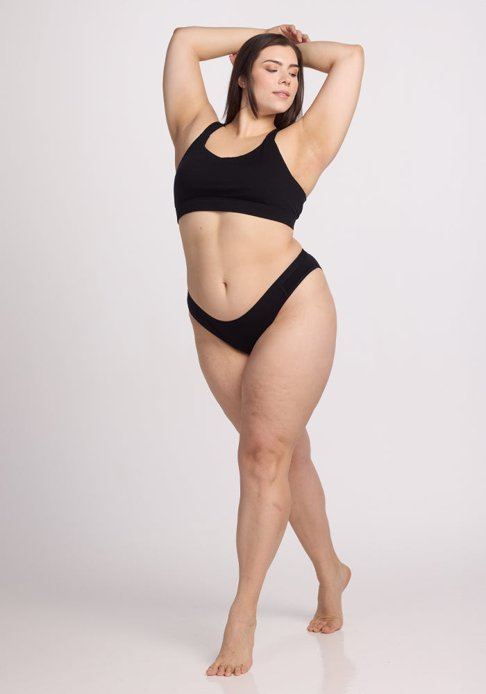 A woman in the Roxie Bikini Underwear - Black by Woolx poses confidently against a plain white background. One arm rests on her hip while the other is raised, highlighting her curves and the smooth lines of her form with full coverage at the back.