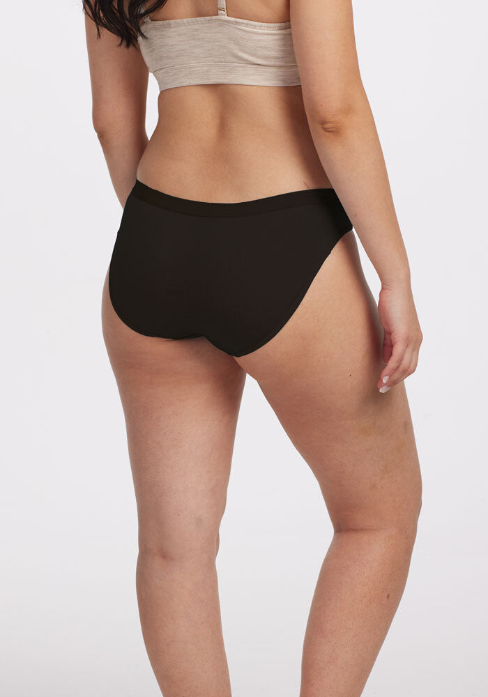 A person is standing with their back to the camera, wearing a beige Merino Wool top and black Roxie Bikini Underwear by Woolx. The fabric appears soft against the plain, light-colored background.