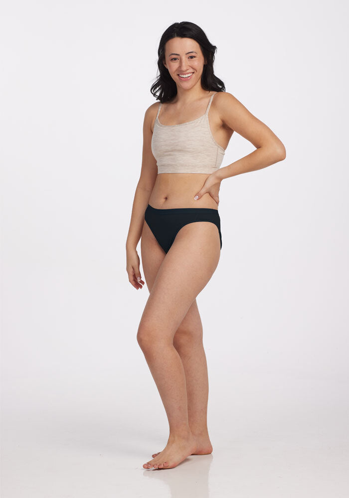 A woman stands against a plain white background, smiling. She is wearing a beige crop top and black Roxie Bikini Underwear by Woolx, both made from feather-light Merino wool. Her left hand rests on her hip while her right arm hangs relaxed by her side. Her dark hair cascades in waves.