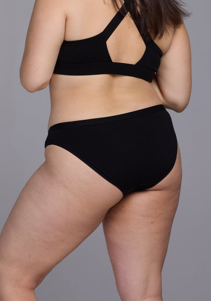 A person wearing the Roxie Bikini Underwear in black by Woolx stands with their back to the camera, showcasing full coverage against a plain gray background.