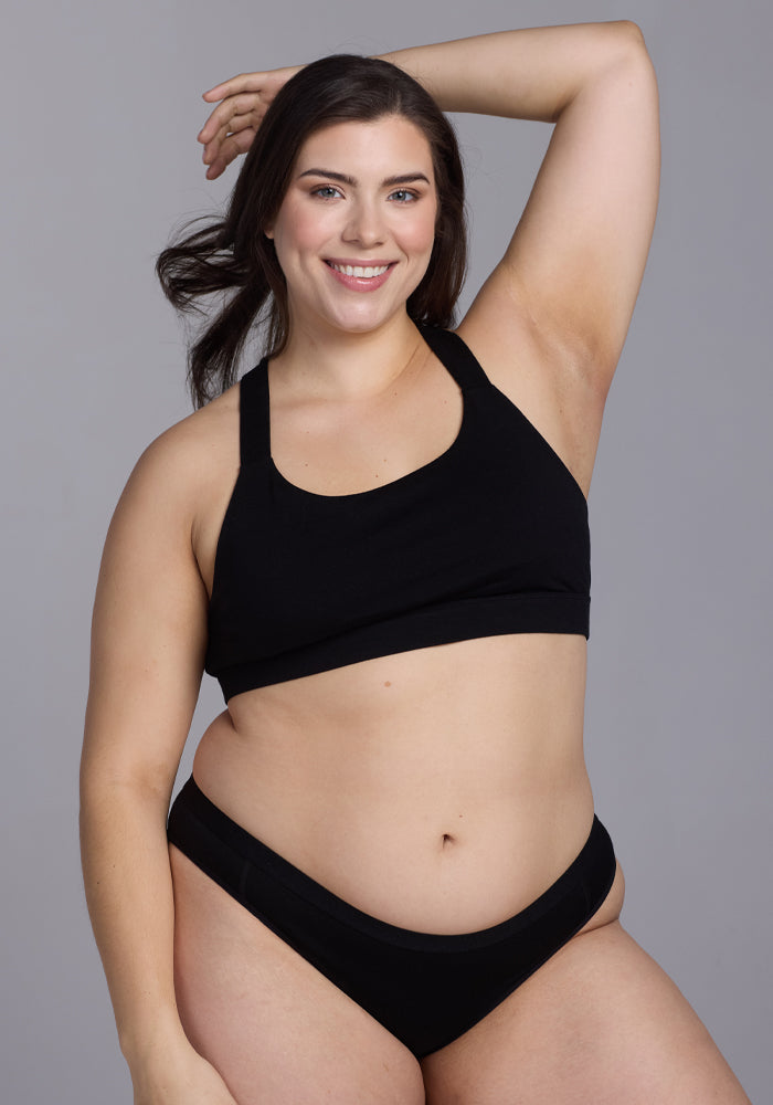 A long-haired woman smiles at the camera with one arm raised, wearing a black sports bra paired with Woolx's Roxie Bikini Underwear in black. The background is plain gray.