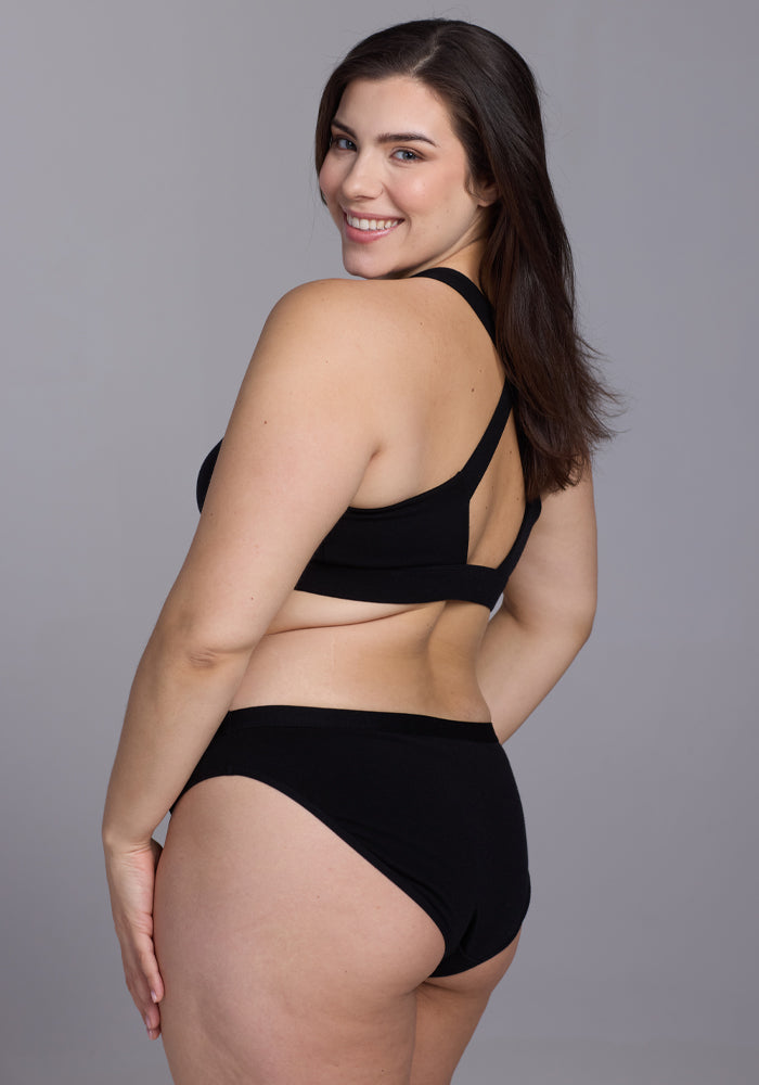 A smiling woman with long dark hair poses against a gray backdrop, wearing Woolx's stylish Roxie Bikini Underwear in black, made from lightweight Merino wool. She glances over her shoulder at the camera, her hand confidently on her hip.