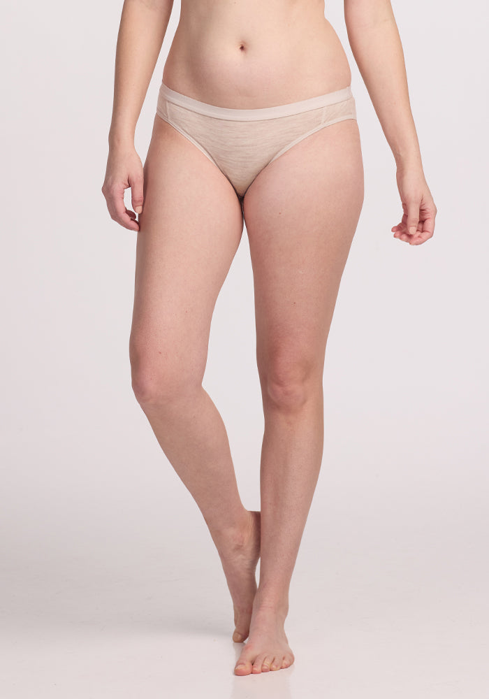 Against a plain backdrop, a person stands in Woolx's Roxie Bikini Underwear in Cream Heather. They're shown waist down, one foot forward for stay-put comfort with every step.