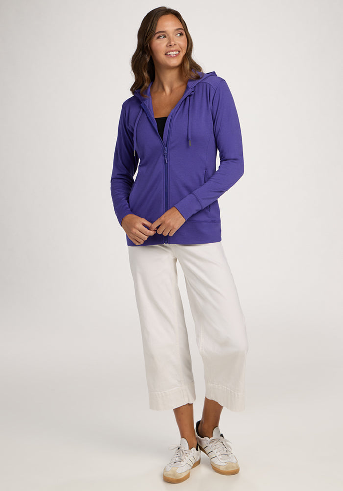 Woman in a Woolx Ryann Hoodie - Berry Indigo, white wide-leg pants, and sneakers, standing and smiling.