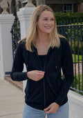 A young woman smiles outdoors in front of a white brick wall and black metal fence, wearing the Woolx Ryann Hoodie in black over a black top and light blue jeans. Her straight blonde hair is loose.